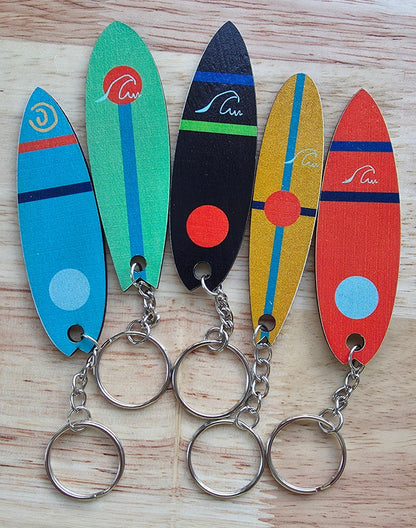Five colorful surfboard-shaped wood keychains with metal split rings, arranged vertically on a light wood background.